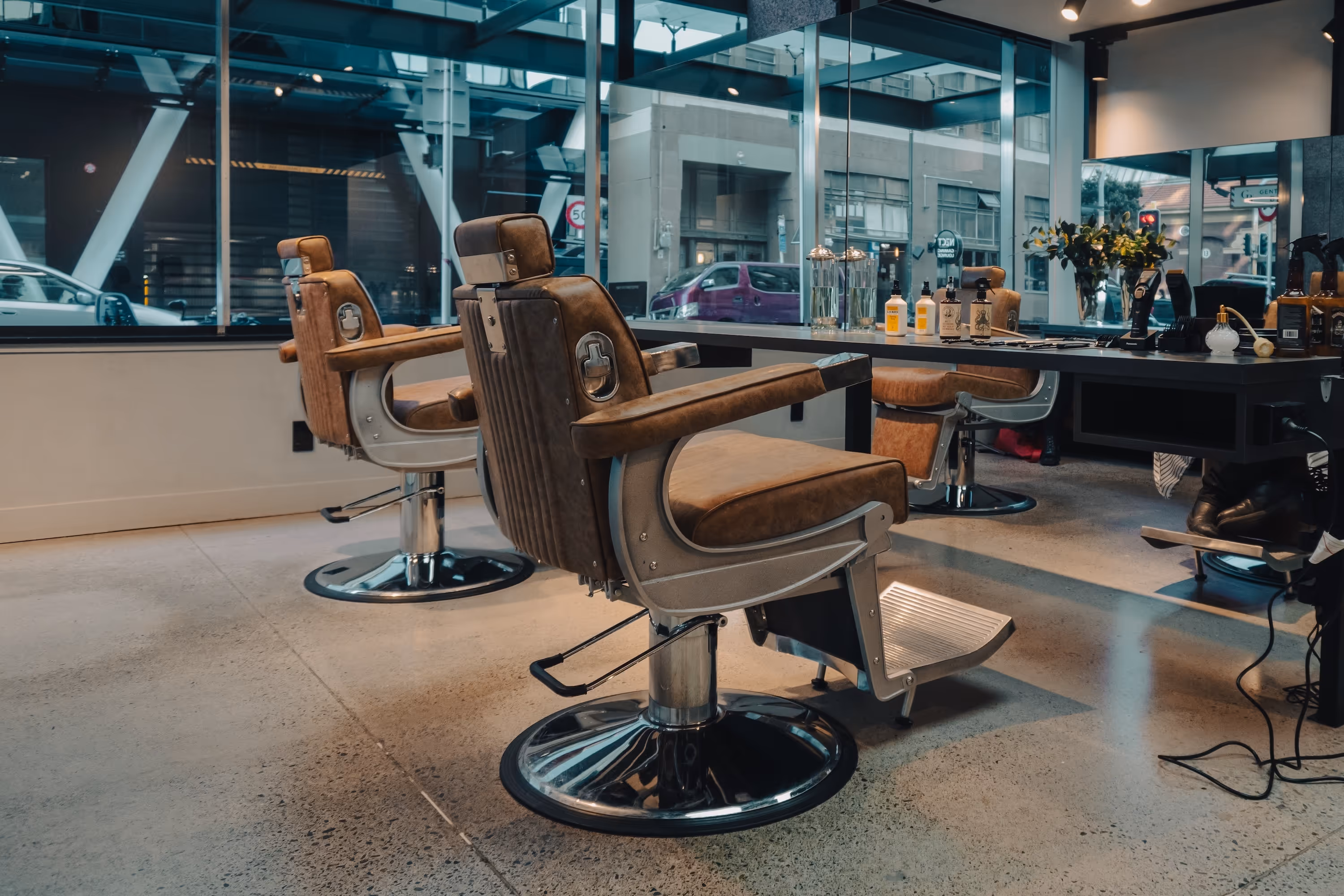 Modern barbershop interior with vintage brown leather barber chairs and grooming products on a counter with a large window view of the street.