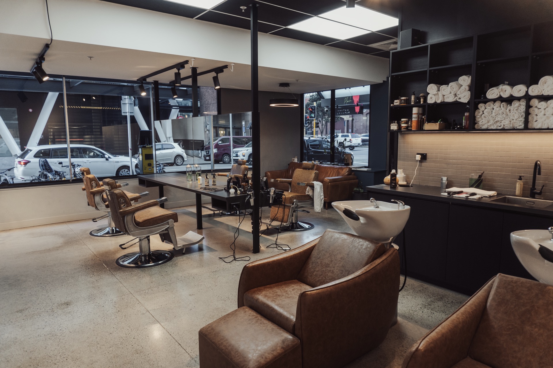 Two barbers styling hair of clients seated in barber chairs in a modern barbershop with wooden floors and skateboard decor.