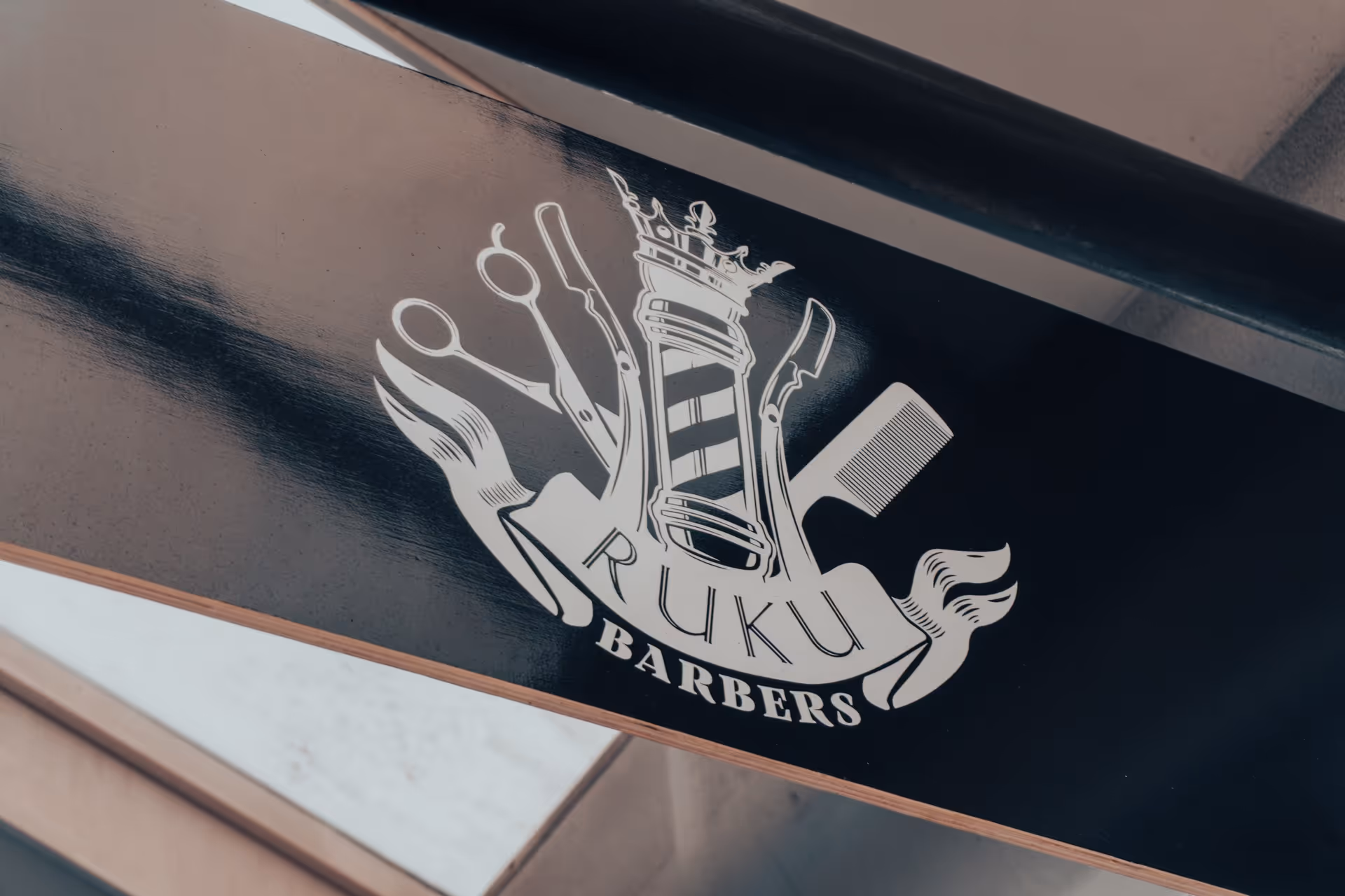 Black wooden sign with a white logo featuring barber tools and a striped barber pole with crown, reading 'RUKU BARBERS'.