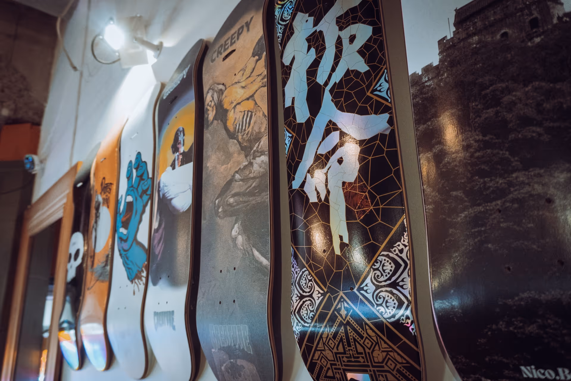 Row of skateboard decks mounted on a wall, each featuring unique artwork including a blue screaming hand, a skeleton labeled 'CREEPY,' and abstract geometric designs.