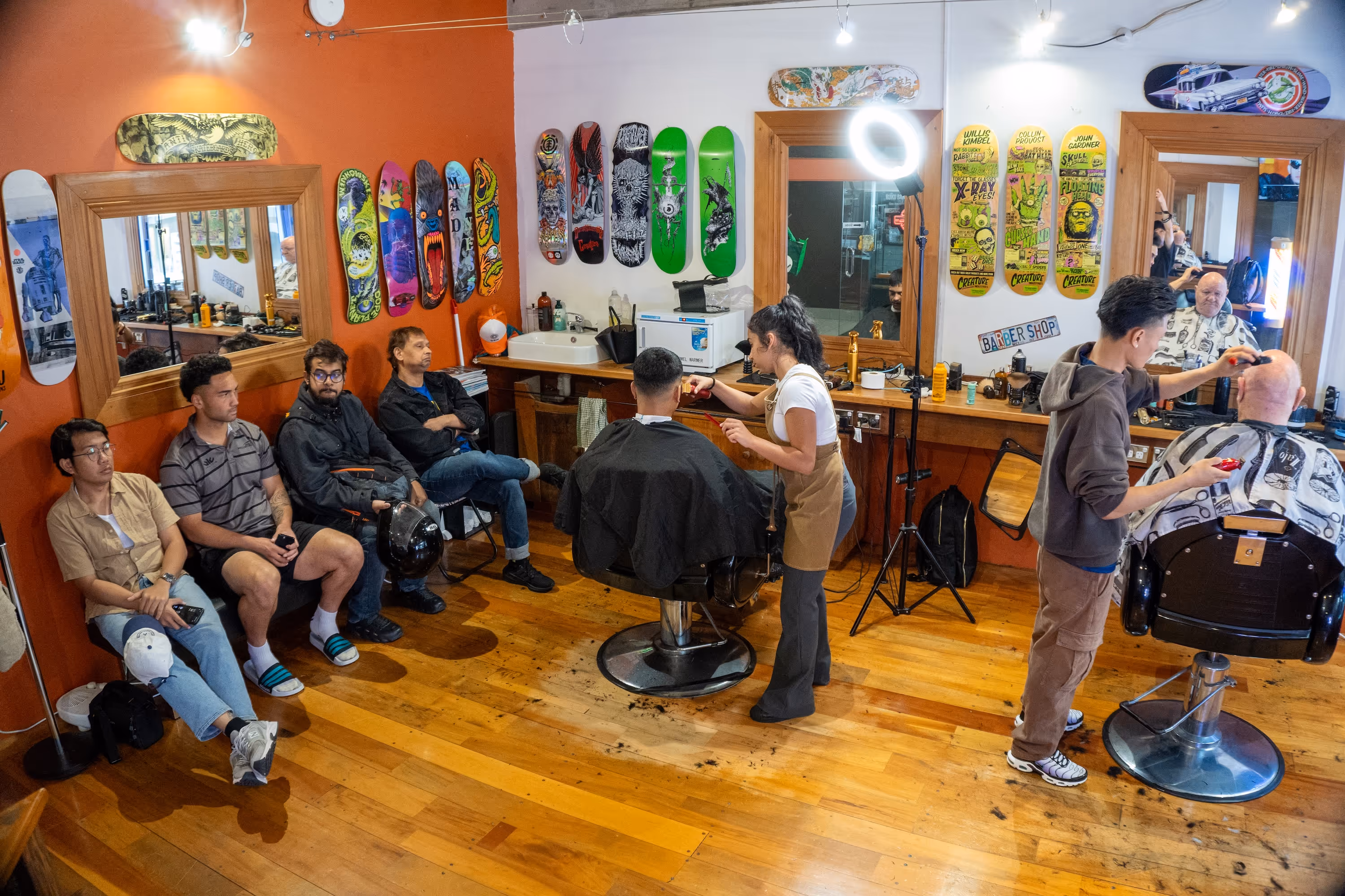 Busy barbershop interior with two barbers cutting hair of seated customers and four men waiting on a bench against a decorated orange wall.