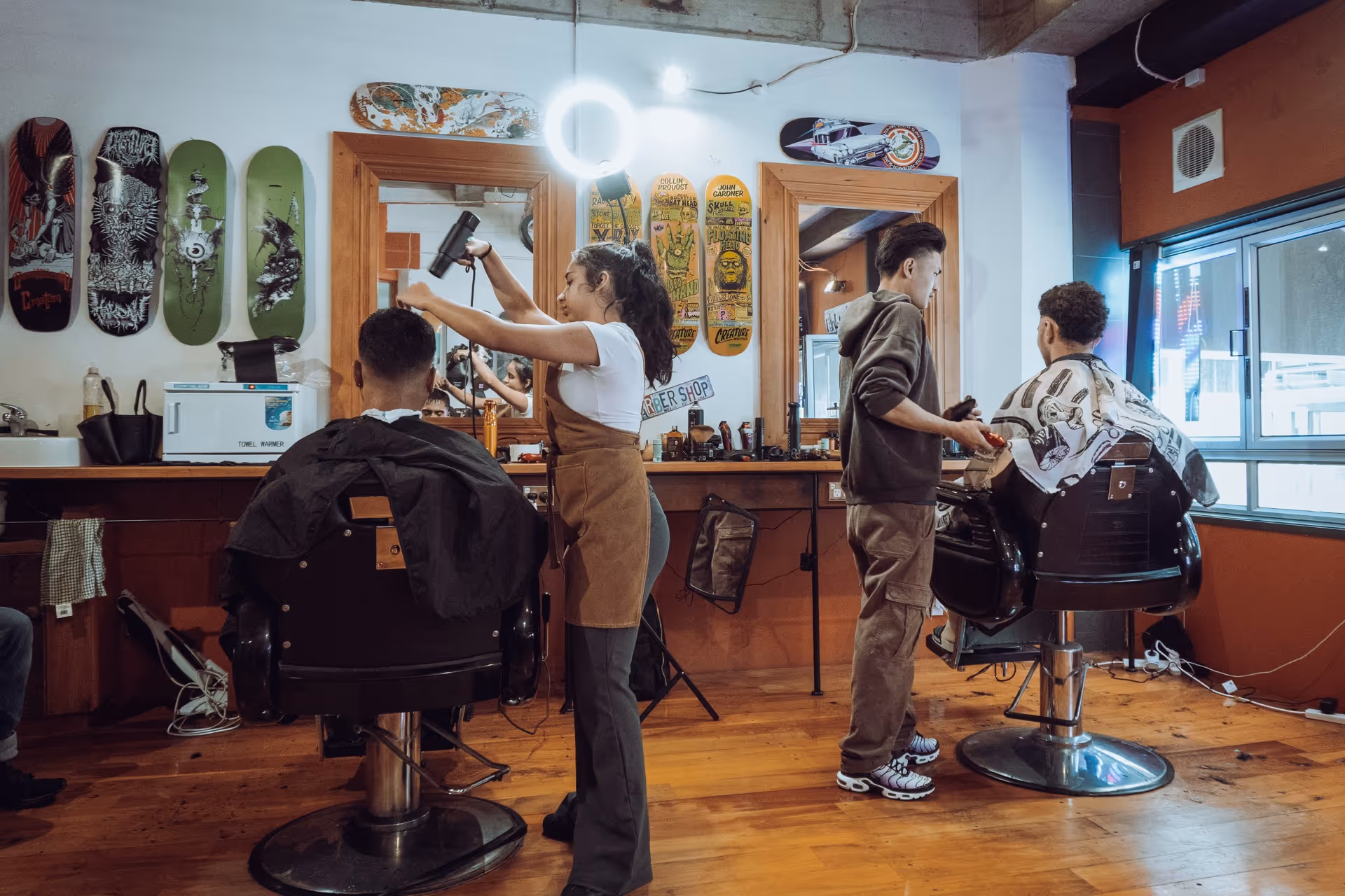 Two barbers styling hair of clients seated in barber chairs in a modern barbershop with wooden floors and skateboard decor.