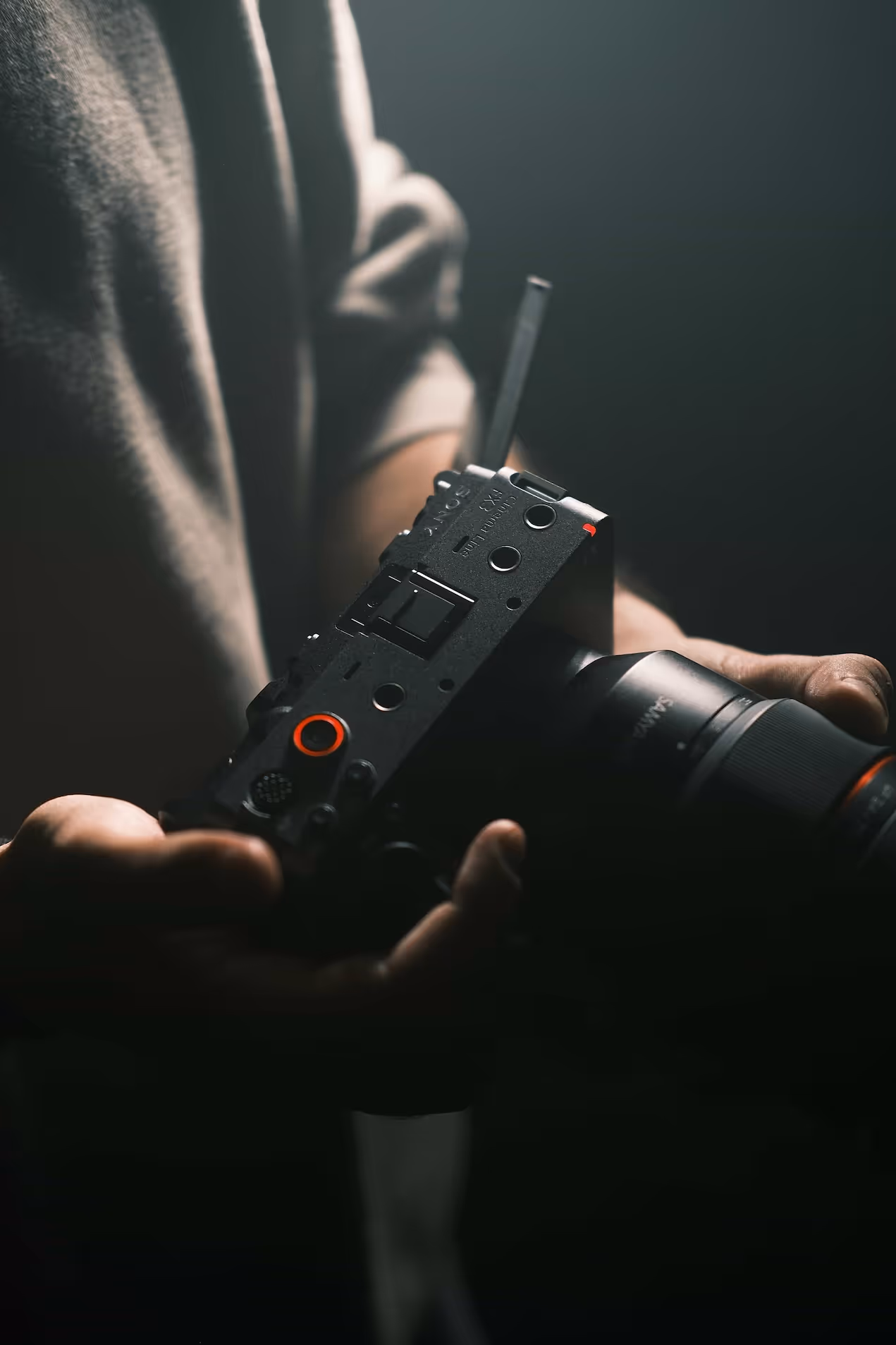 A close-up of a person's hands holding a black camera rig with orange accents, against a dark, blurred background. The focus is on the rig's controls and structure.